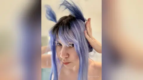 Young woman with long lavender blue hair styling her bangs in a bright indoor setting.