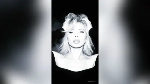 SerenaStar poses with her hands in her blonde hair, wearing a black off-the-shoulder top against a dark background.