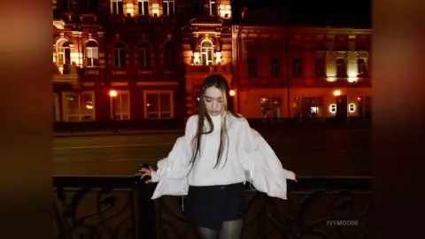 Young woman in a white sweater and dark skirt standing by a railing in front of a lit red building at night.