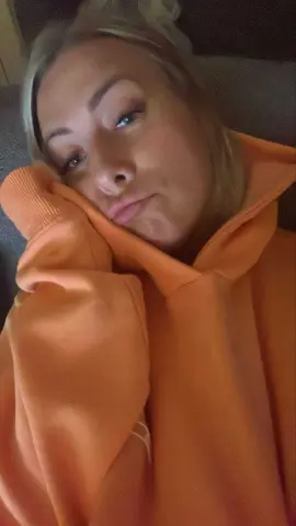 Blonde woman in a cozy orange hoodie relaxing indoors with a calm expression.