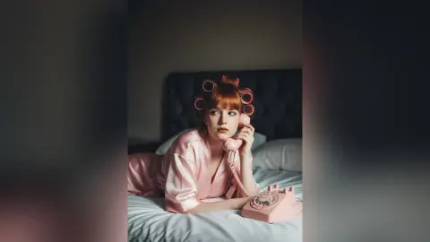 Redheaded woman in pink silk pajamas with hair rollers talking on a vintage pink telephone in a moody bedroom.