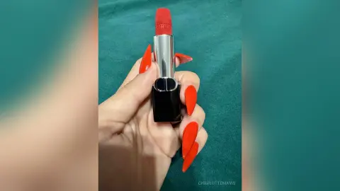 Hand with long red manicured nails holding a red lipstick tube against a teal background.