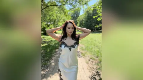 Elegant woman in a white satin lace slip dress walking through a sunny green park.