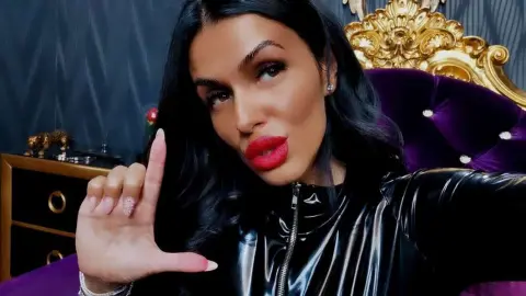 Glamorous woman in a black shiny latex outfit posing with red lips in a luxurious purple velvet bedroom.