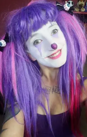 Woman in purple clown wig and white face paint smiling in a playful cosplay costume.