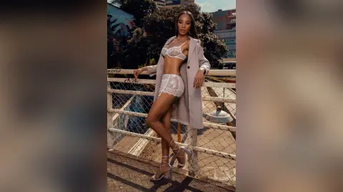 Stylish woman in white lace lingerie and a long beige coat posing on a sunny urban balcony.
