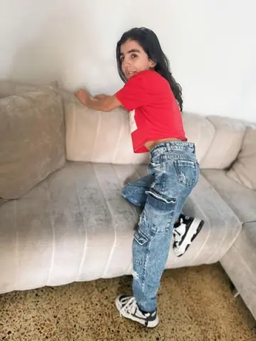 Young woman in a red t-shirt and baggy blue jeans posing playfully on a beige sofa in a bright room.