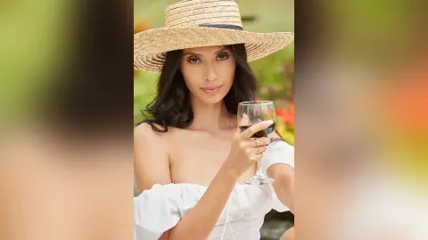 LucyMartin wears a wide-brimmed straw hat and a white off-the-shoulder top while holding a glass of red wine.