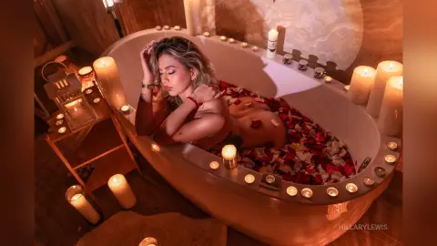 Relaxed woman in a rose petal bath surrounded by glowing candles in a cozy, romantic candlelit setting.