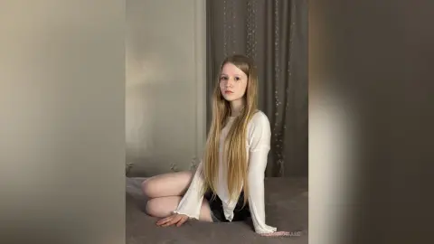 Blonde woman in a white long-sleeve shirt sitting on a bed in a minimalist, moody room.