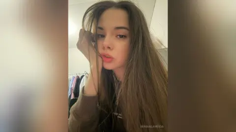 Young woman with long brown hair wearing a casual dark sweatshirt in a soft, moody indoor setting.