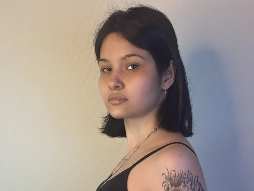 JacquiKozma poses with a direct gaze, showing her short black hair and shoulder tattoo against a plain wall.
