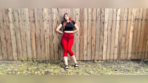 Young woman in a black t-shirt and red trousers leaning against a rustic wooden fence in a casual outdoor setting.
