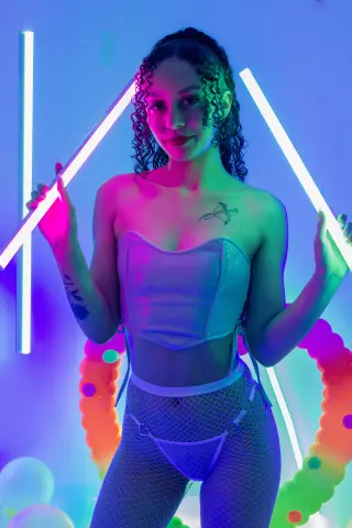 Young woman in white corset and fishnet tights posing under neon lights in a vibrant, colorful studio setting.