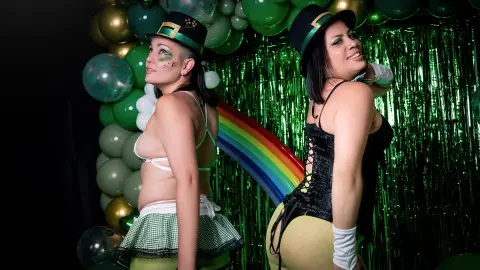 Two smiling women in festive green outfits and hats posing before a rainbow and green balloon backdrop.