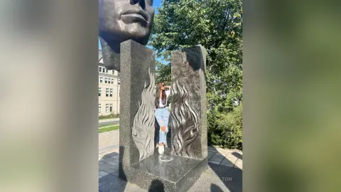 Woman in white shirt and jeans standing by a large stone face sculpture in a sunny outdoor plaza.
