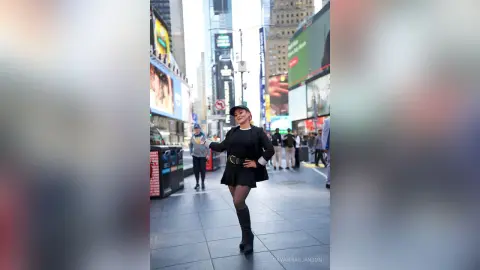 Stylish woman in a black dress and boots smiling on a busy New York City street near Times Square billboards.