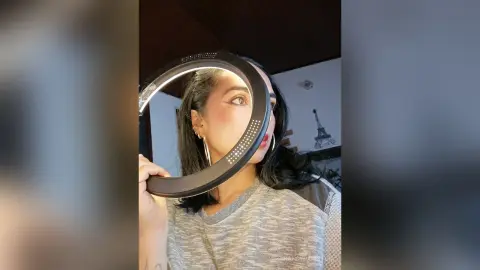 Young woman in a grey sweater holding a circular ring light for a cozy indoor selfie.