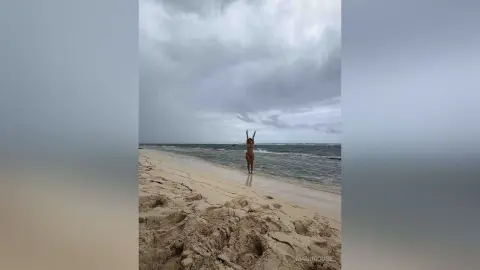 Joyful woman in a bikini standing on a sandy beach under a dramatic cloudy sky.
