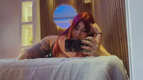 Red-haired elf cosplayer in dark outfit taking a mirror selfie in a modern bedroom setting.