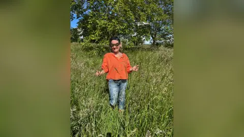 Smiling woman in an orange shirt and jeans standing in a sunny green meadow under leafy trees.
