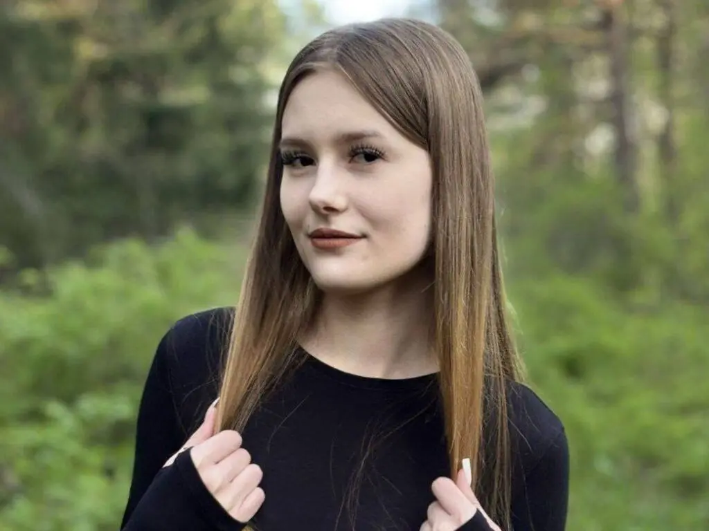 Young woman with long straight hair wearing a black long-sleeve shirt in a lush green forest setting.