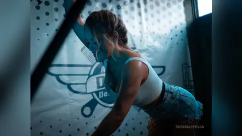 Young woman in a white tank top and denim shorts posing against a branded backdrop in moody blue lighting.