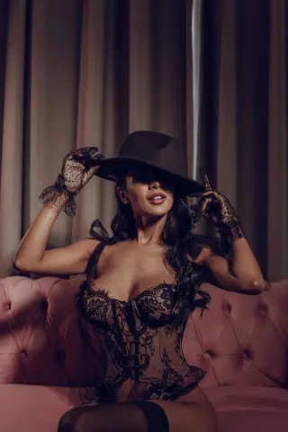 AnnyHoney poses on a pink sofa wearing a black lace corset and a wide-brimmed black hat.
