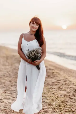Smiling woman in a white linen sundress holding a wildflower bouquet on a sunny beach at sunset.
