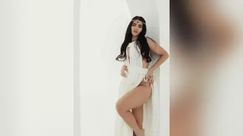 Elegant woman in a flowing white Grecian dress posing against a bright white wall in a dreamy, ethereal mood.