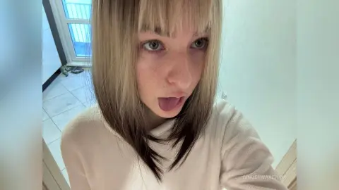 Blonde woman with bangs in a white sweatshirt making a playful expression in a bright room.