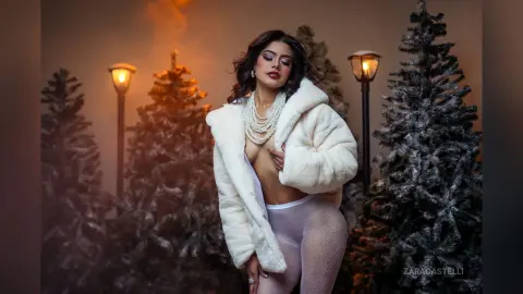 Glamorous woman in a white faux fur coat and tights posing amidst snowy pine trees under warm lantern light.