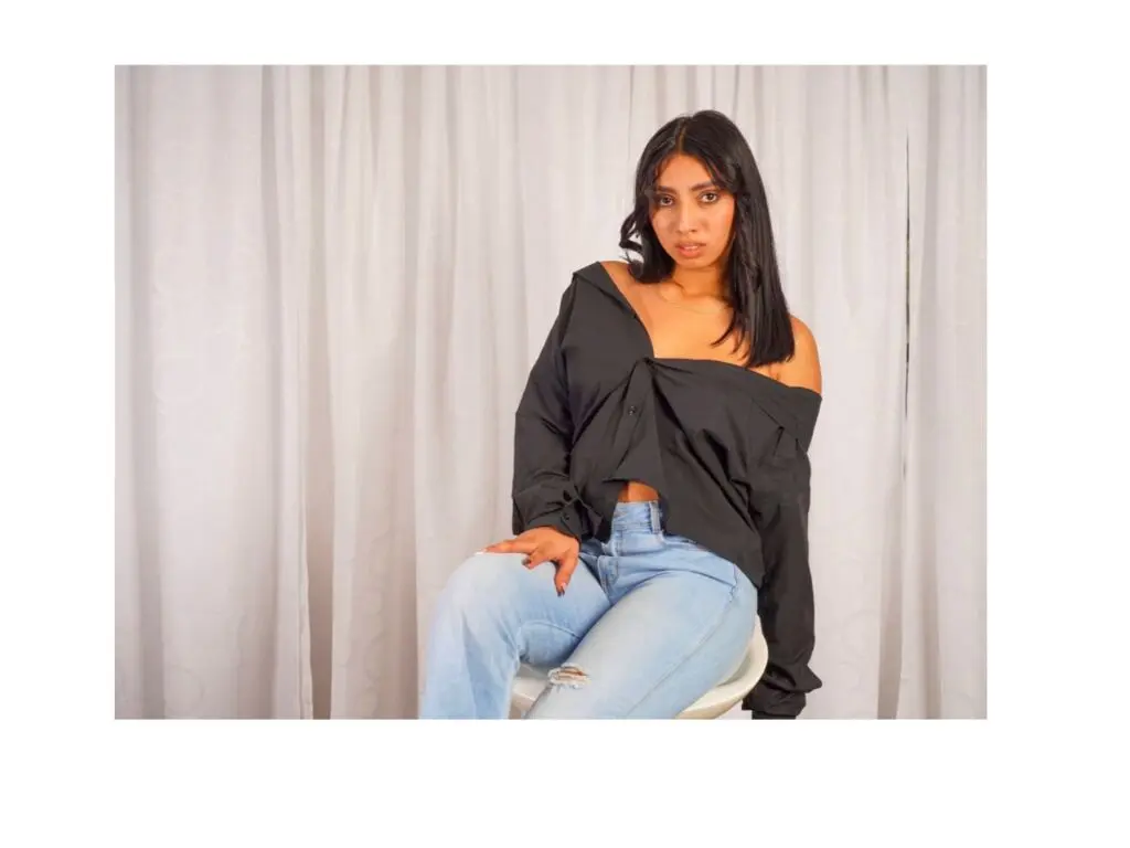Young woman in a black off-the-shoulder blouse and light blue jeans sitting against a white curtain backdrop.