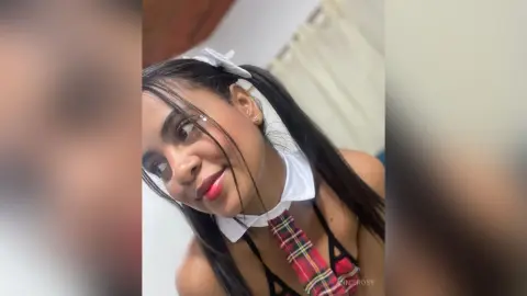 AnnieRosy smiles while wearing a white collar and plaid schoolgirl outfit with her long black hair styled in pigtails.