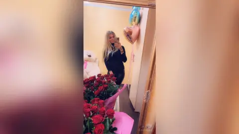 Blonde woman in black outfit taking a selfie near red roses and heart balloons for a romantic celebration.