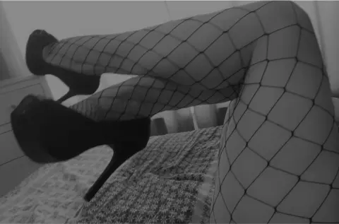 Woman in fishnet tights and black high heels relaxing on a bed in a moody black and white aesthetic.