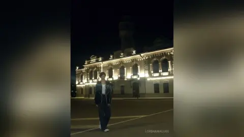 Young man in a dark jacket and jeans standing in front of a lit classical building at night.