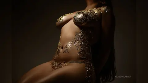Artistic portrait of a woman with shimmering gold body paint in a moody, dark studio setting.