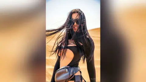 Stylish woman with long dark hair in a black bodysuit and fanny pack posing in a sunny desert landscape.