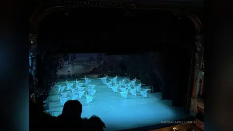 Ballet dancers in white tutus performing a graceful classical dance on a moonlit stage during a theater production.