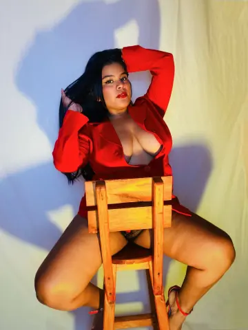 Curvy woman in a red blazer sitting on a wooden chair against a white backdrop in a moody studio setting.