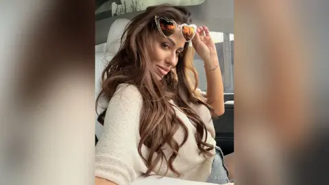 Smiling woman with long wavy hair wearing a beige sweater and heart-shaped sunglasses in a car interior.