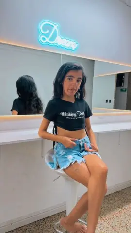 Young woman in black graphic t-shirt and denim shorts sitting in a modern salon with neon Dream sign.