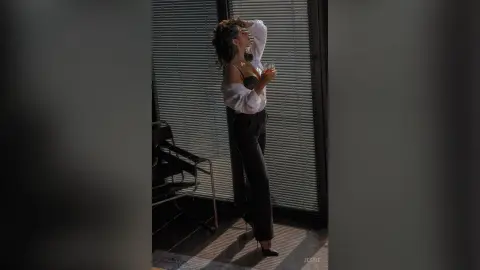 Elegant woman in a white blouse and black trousers holding a drink by a window in moody sunlight.