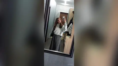 Smiling woman in a white sweater and patterned maxi skirt taking a mirror selfie in a modern dressing room.