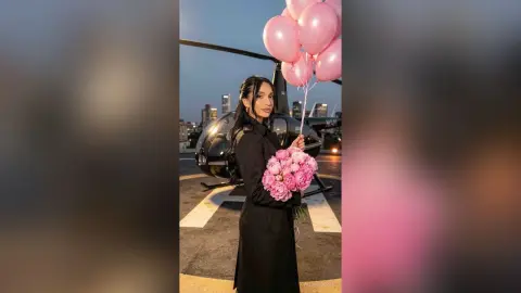 Elegant woman in a black coat holding pink flowers and balloons near a helicopter at sunset.