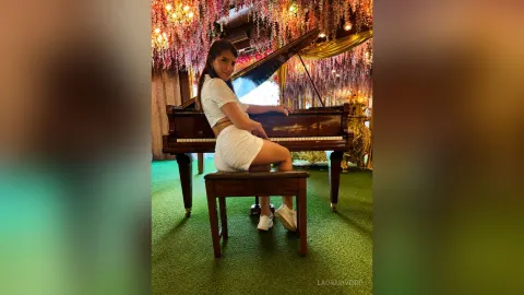 LauraRiveiro — LauraRiveiro sits on a piano bench wearing a white top and skirt in a room filled with flowers.