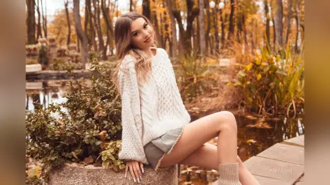 Smiling young woman in a cozy white knit sweater sitting by a pond in an autumnal forest setting.