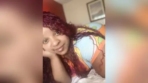 Smiling woman with long curly red hair wearing a colorful patterned top in a cozy indoor setting.