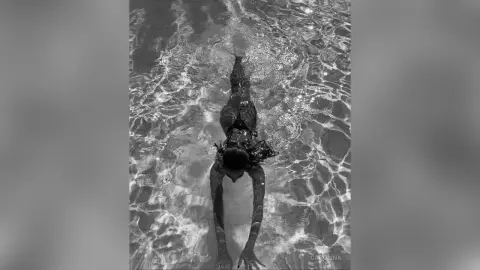 Black and white overhead view of a woman swimming in clear rippling water wearing a dark swimsuit.
